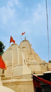 The image features a traditional Indian temple with intricately carved spires and domes, adorned with orange flags. The temple is painted in a light cream color against a clear blue sky.