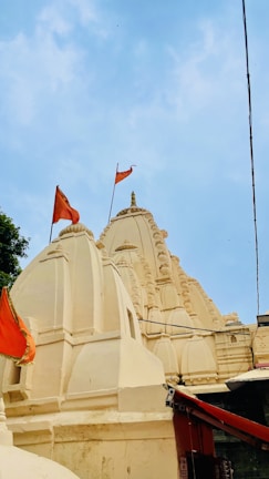 The image features a traditional Indian temple with intricately carved spires and domes, adorned with orange flags. The temple is painted in a light cream color against a clear blue sky.