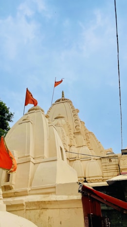 The image features a traditional Indian temple with intricately carved spires and domes, adorned with orange flags. The temple is painted in a light cream color against a clear blue sky.