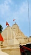 The image features a traditional Indian temple with intricately carved spires and domes, adorned with orange flags. The temple is painted in a light cream color against a clear blue sky.
