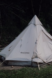 A white Naturehike brand tent is set up in a natural outdoor setting with surrounding greenery. The tent has a triangular, teepee-like shape and is partially shaded by trees. The ground in front of the tent consists of grass and some dirt.