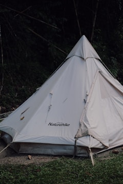 A white Naturehike brand tent is set up in a natural outdoor setting with surrounding greenery. The tent has a triangular, teepee-like shape and is partially shaded by trees. The ground in front of the tent consists of grass and some dirt.