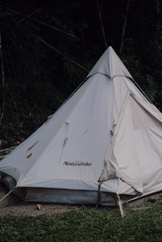 A white Naturehike brand tent is set up in a natural outdoor setting with surrounding greenery. The tent has a triangular, teepee-like shape and is partially shaded by trees. The ground in front of the tent consists of grass and some dirt.
