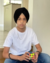 A young person wearing a black turban and a white t-shirt sits holding a Rubik's Cube. A necklace and a bracelet are visible, and the background consists of a neutral-colored interior with soft lighting.