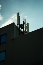 A rooftop with multiple telecommunications antennas and equipment is silhouetted against a clear blue sky. There are minimal details from the building, with emphasis on the antennas reaching upwards. Scattered clouds add a soft texture to the sky, enhancing the contrast with the foreground structure.