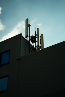 A rooftop with multiple telecommunications antennas and equipment is silhouetted against a clear blue sky. There are minimal details from the building, with emphasis on the antennas reaching upwards. Scattered clouds add a soft texture to the sky, enhancing the contrast with the foreground structure.