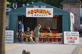 A green tent with a sign reading 'Aboriginal Embassy' is placed in an outdoor area. Inside, several people sit on chairs facing a screen. Two prominent signs outside the tent express political messages about indigenous rights and land ownership.