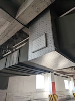 An industrial setting with large metal ducts and pipes running along the ceiling. The ducts are metallic with a diamond-shaped pattern. There are vents and some panels, with supporting structures attached to the roof. The floor area has a column painted white with an orange stripe and black-and-yellow hazard markings at the base. Natural light is coming in through high windows on the wall.