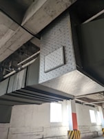 An industrial setting with large metal ducts and pipes running along the ceiling. The ducts are metallic with a diamond-shaped pattern. There are vents and some panels, with supporting structures attached to the roof. The floor area has a column painted white with an orange stripe and black-and-yellow hazard markings at the base. Natural light is coming in through high windows on the wall.