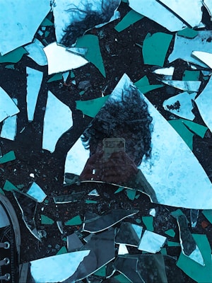 A reflection of a person with curly hair is visible in shards of broken mirror scattered on a dark surface. The fragments create a fragmented and distorted image with pieces of the sky and surrounding environment. A shoe is partially visible in the bottom left corner.