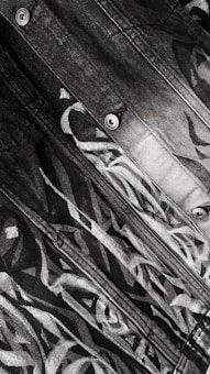 Close-up view of a denim fabric with an abstract, monochromatic design. The pattern consists of intertwining lines and shapes with a gradient transition from light to dark. Visible metal buttons are part of the fabric.