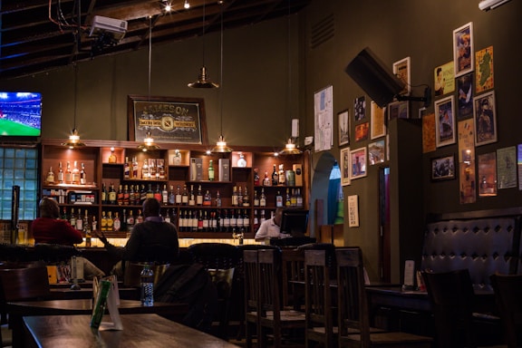 A cozy bar interior with dim lighting and a variety of bottles displayed on shelves behind the counter. Several people are seated at the bar, and the walls are adorned with numerous framed pictures and posters. A television screen in the corner shows a sports game. The atmosphere is intimate, with warm tones from the wooden furniture and soft lighting from the hanging lamps.