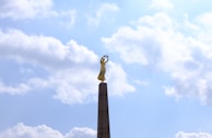A tall monument topped with a golden statue stands against a bright sky filled with scattered white clouds. The statue is elegantly poised, contributing to a sense of grandeur and majesty.