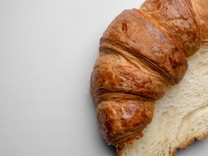A close-up of a flaky, golden-brown croissant partially sliced, showcasing its airy and layered interior against a neutral background.