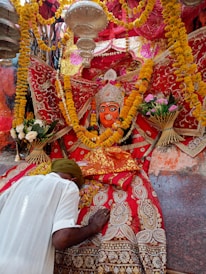 A vibrant religious scene features a richly adorned deity statue surrounded by colorful garlands and intricate fabrics. A person bows down in reverence, placing their head and hands on a decorated surface. The atmosphere is festive and spiritual, with yellow and orange marigold flowers prominently displayed.