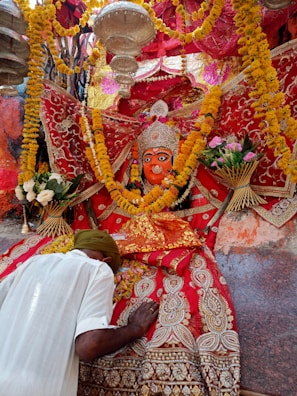 A vibrant religious scene features a richly adorned deity statue surrounded by colorful garlands and intricate fabrics. A person bows down in reverence, placing their head and hands on a decorated surface. The atmosphere is festive and spiritual, with yellow and orange marigold flowers prominently displayed.