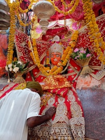 A vibrant religious scene features a richly adorned deity statue surrounded by colorful garlands and intricate fabrics. A person bows down in reverence, placing their head and hands on a decorated surface. The atmosphere is festive and spiritual, with yellow and orange marigold flowers prominently displayed.
