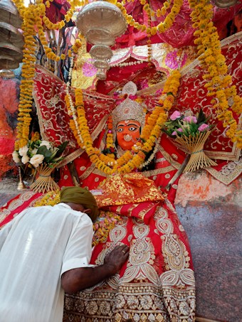 A vibrant religious scene features a richly adorned deity statue surrounded by colorful garlands and intricate fabrics. A person bows down in reverence, placing their head and hands on a decorated surface. The atmosphere is festive and spiritual, with yellow and orange marigold flowers prominently displayed.