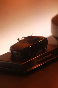A detailed model of a car, specifically a Honda S2000 Roadster, is placed on a pedestal or stand. The lighting creates a warm, dramatic effect that highlights the features of the car model.