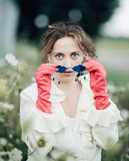 A person with expressive eyes is seen holding up a whimsical accessory shaped like a mustache with dangling blue teardrop beads. The individual is wearing bright pink gloves and is surrounded by a natural environment with soft-focus vegetation, giving a playful and surreal impression.