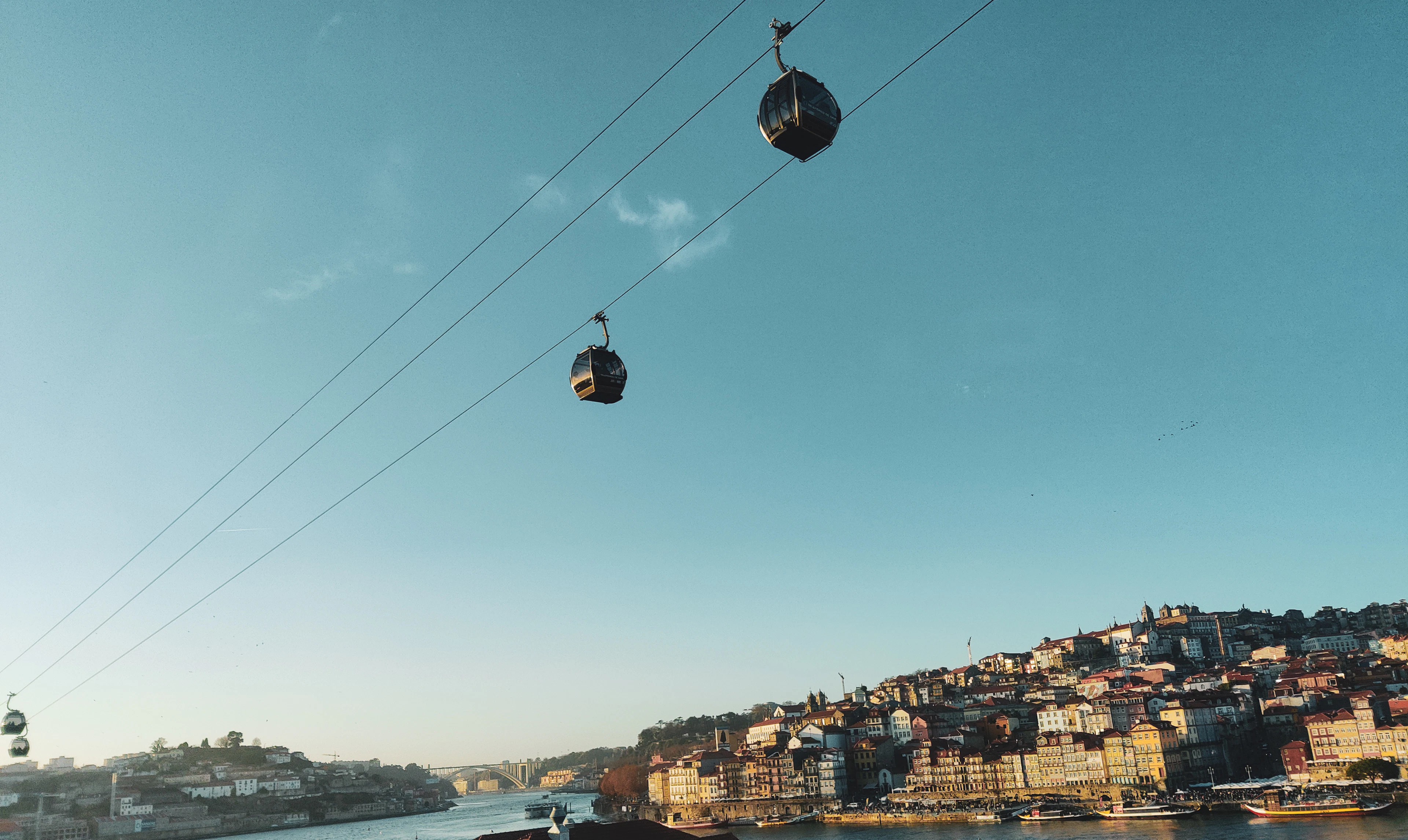 Two gondolas of the Gaia cable car hanging from their wire high over the Douro, with the sunlit tiers of Porto's riverfront houses and a bridge in the distance below.
