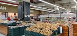A supermarket produce section featuring a variety of vegetables and fruits displayed in baskets and crates. Various tubers, pumpkins, and other fresh items are visible. The store has a spacious interior with overhead lights and white ceilings. Shelves filled with products and signs are in the background.