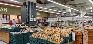 A supermarket produce section featuring a variety of vegetables and fruits displayed in baskets and crates. Various tubers, pumpkins, and other fresh items are visible. The store has a spacious interior with overhead lights and white ceilings. Shelves filled with products and signs are in the background.