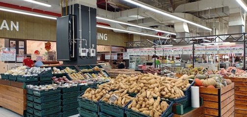 A supermarket produce section featuring a variety of vegetables and fruits displayed in baskets and crates. Various tubers, pumpkins, and other fresh items are visible. The store has a spacious interior with overhead lights and white ceilings. Shelves filled with products and signs are in the background.