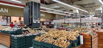 A supermarket produce section featuring a variety of vegetables and fruits displayed in baskets and crates. Various tubers, pumpkins, and other fresh items are visible. The store has a spacious interior with overhead lights and white ceilings. Shelves filled with products and signs are in the background.