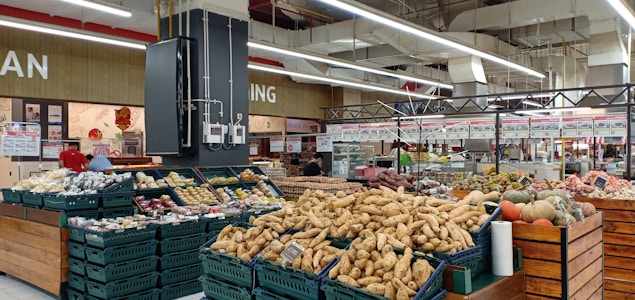 A supermarket produce section featuring a variety of vegetables and fruits displayed in baskets and crates. Various tubers, pumpkins, and other fresh items are visible. The store has a spacious interior with overhead lights and white ceilings. Shelves filled with products and signs are in the background.