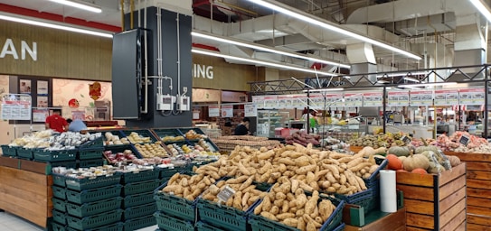 A supermarket produce section featuring a variety of vegetables and fruits displayed in baskets and crates. Various tubers, pumpkins, and other fresh items are visible. The store has a spacious interior with overhead lights and white ceilings. Shelves filled with products and signs are in the background.