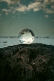 A clear crystal ball is placed on a rugged, dark rock surface, reflecting the sky and clouds above, with a distant horizon visible in the background. The scene conveys a sense of tranquility and mystique.