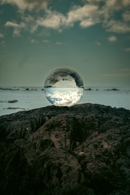 A clear crystal ball is placed on a rugged, dark rock surface, reflecting the sky and clouds above, with a distant horizon visible in the background. The scene conveys a sense of tranquility and mystique.