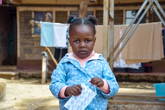 A young child stands in an outdoor setting, holding a face mask and wearing a blue patterned jacket. In the background, there are hanging clotheslines with laundry and a building with wooden and metal structures.