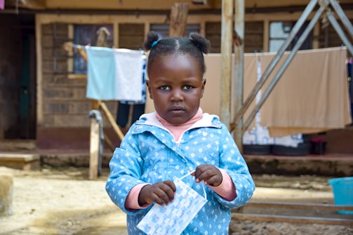 A young child stands in an outdoor setting, holding a face mask and wearing a blue patterned jacket. In the background, there are hanging clotheslines with laundry and a building with wooden and metal structures.