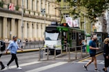 A modern tram is stopped at a station in a busy urban environment. The street is lined with historic buildings, and various pedestrians are walking, some appearing engaged with their phones. The tram is green and white, with a destination sign displaying the route number. Leaves from nearby trees scatter the edges of the street.