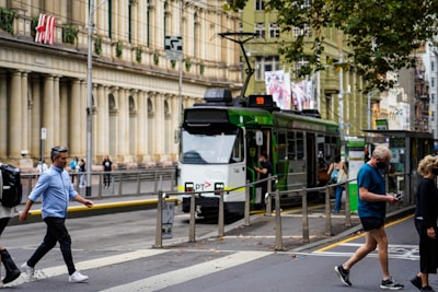 A modern tram is stopped at a station in a busy urban environment. The street is lined with historic buildings, and various pedestrians are walking, some appearing engaged with their phones. The tram is green and white, with a destination sign displaying the route number. Leaves from nearby trees scatter the edges of the street.