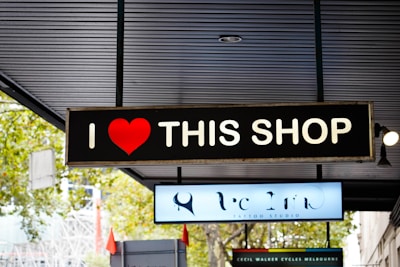 A black rectangular sign with white text and a red heart symbol, reading 'I ❤️ THIS SHOP'. The sign is mounted under a ceiling with visible lights and architecture. In the background, other shop signs and some greenery are partially visible.