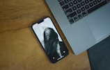 A smartphone with a black and white lock screen image of a person partially obscured by their hair is lying on a wooden surface beside a laptop keyboard.