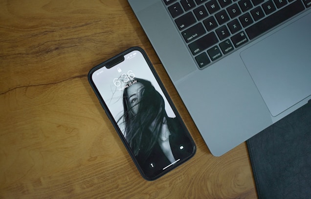 A smartphone with a black and white lock screen image of a person partially obscured by their hair is lying on a wooden surface beside a laptop keyboard.