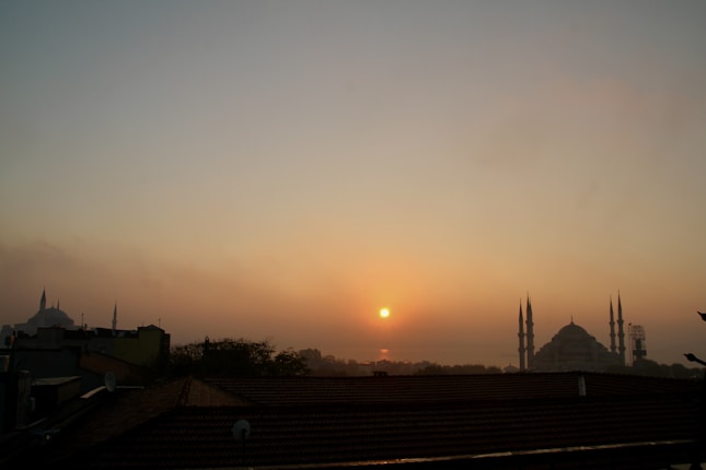 A serene sunrise over a cityscape featuring two prominent mosques with minarets silhouetted against an orange and pink sky. The rooftops and trees in the foreground add depth to the composition.