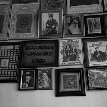 A collection of framed portraits and calligraphy hanging on a wall. The frames vary in size and style, with some displaying detailed calligraphic art in what appears to be Arabic script, while others contain black and white photographs of elderly men in traditional attire. The arrangement is dense, creating a collage-like effect.