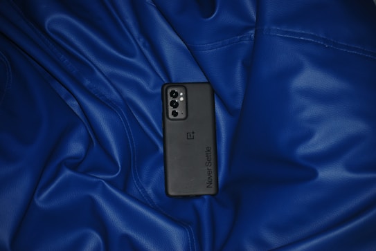 A smartphone with a sleek back design and the words 'Never Settle' is placed on rumpled blue fabric. The phone has a triple-camera setup and a flash.