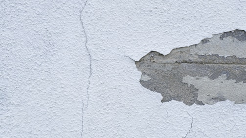 A textured wall with a noticeable crack running from top to bottom, alongside a large area where the plaster is peeling away to reveal the underlying concrete.