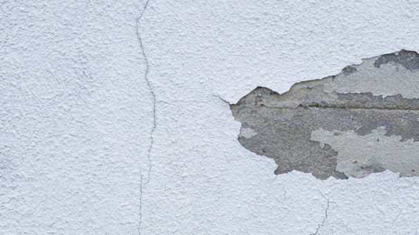 A textured wall with a noticeable crack running from top to bottom, alongside a large area where the plaster is peeling away to reveal the underlying concrete.