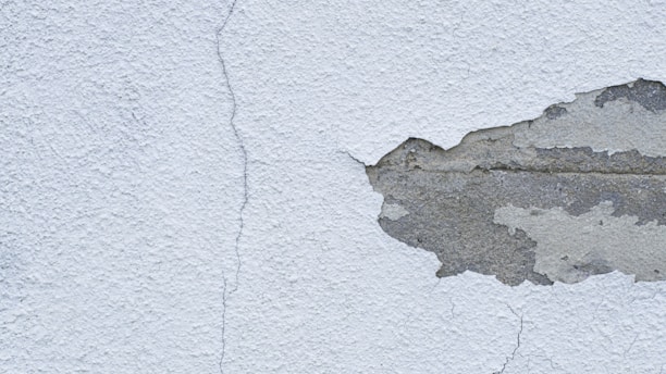 A textured wall with a noticeable crack running from top to bottom, alongside a large area where the plaster is peeling away to reveal the underlying concrete.