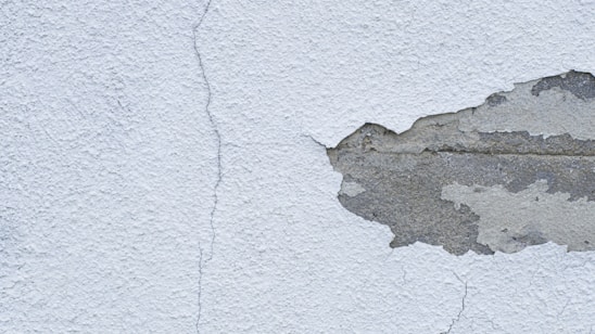 A textured wall with a noticeable crack running from top to bottom, alongside a large area where the plaster is peeling away to reveal the underlying concrete.