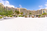 A beachfront resort with a large, elegant building featuring multiple stories and balconies, surrounded by palm trees. The foreground consists of a white sandy beach with scattered lounge chairs and umbrellas. The sky is clear with a few clouds, indicating a sunny day.