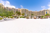 A beachfront resort with a large, elegant building featuring multiple stories and balconies, surrounded by palm trees. The foreground consists of a white sandy beach with scattered lounge chairs and umbrellas. The sky is clear with a few clouds, indicating a sunny day.