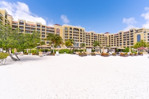 A beachfront resort with a large, elegant building featuring multiple stories and balconies, surrounded by palm trees. The foreground consists of a white sandy beach with scattered lounge chairs and umbrellas. The sky is clear with a few clouds, indicating a sunny day.